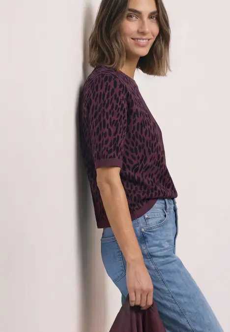 Frau lehnt lächelnd an der Wand, trägt lila Leoparden-Pullover und blaue Jeans.