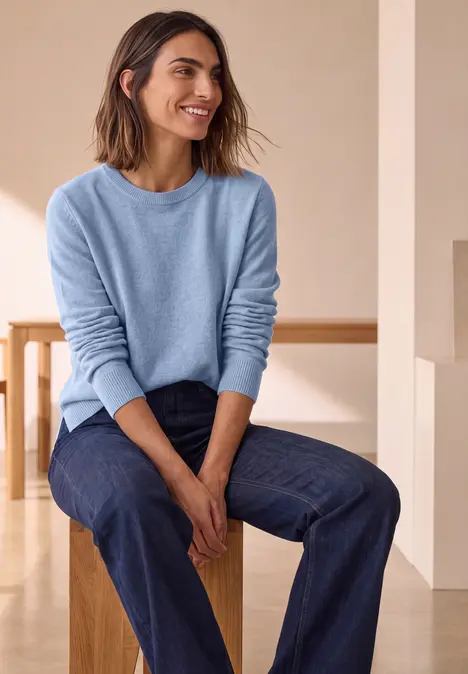 Frau mit blauem Pullover und Jeans sitzt lächelnd auf Hocker in hellem Raum.