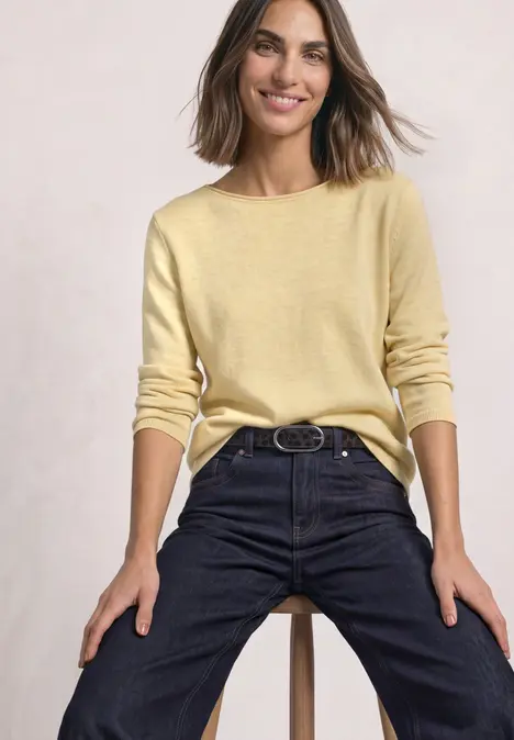 Eine lächelnde Frau sitzt auf einem Hocker, trägt gelben Pullover und dunkle Jeans.