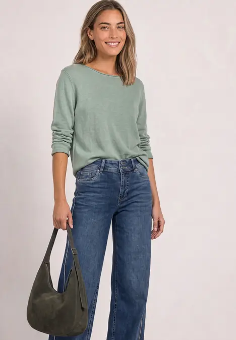 Eine Frau mit graugrünem Langarmshirt, blauen Jeans und grüner Schultertasche, die lächelnd posiert.
