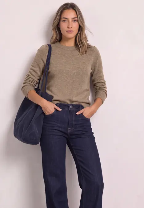 Vrouw in beige trui en donkere jeans met blauwe schoudertas voor lichte achtergrond.