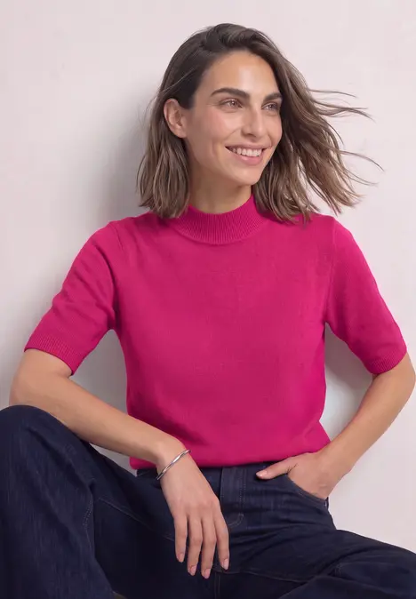Een glimlachende vrouw met kort bruin haar, gekleed in een felroze korte mouw top en een spijkerbroek.