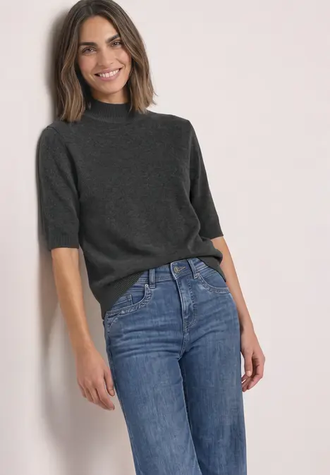 Femme souriante vêtue d'un pull gris foncé à manches courtes et d'un jean bleu, appuyée sur un mur clair.