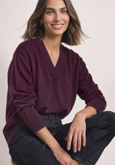 Eine lächelnde Frau im violetten V-Pullover und schwarzen Jeans sitzt entspannt.