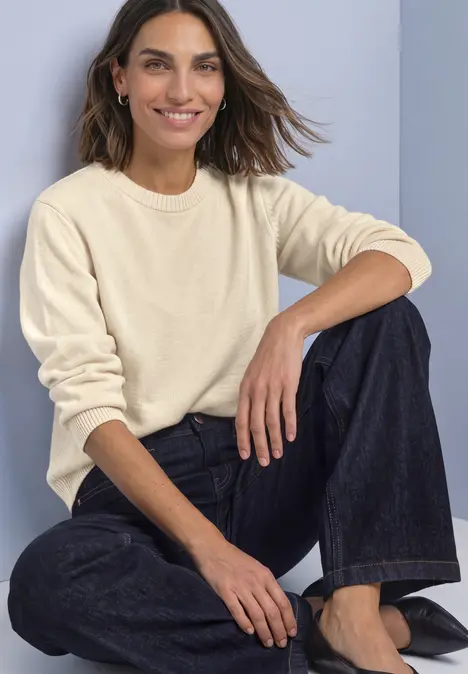 Frau in cremefarbenem Pullover und Jeans sitzt lächelnd auf dem Boden.