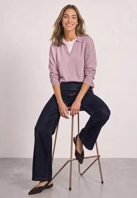 Femme souriante assise sur un tabouret, porte un pull rose col V et un jean large.