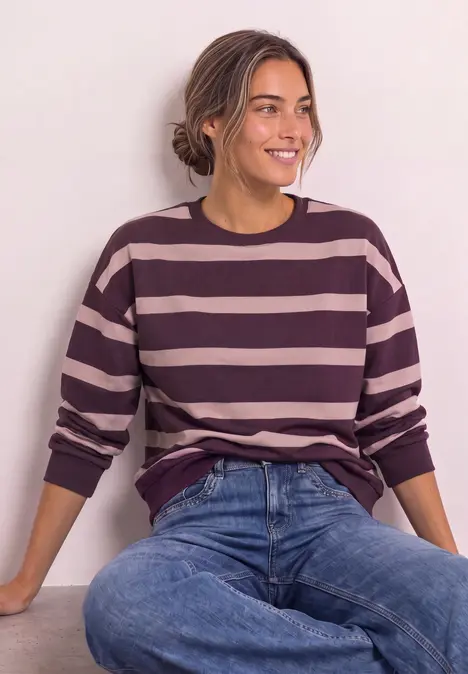 Frau in gestreiftem Pullover (rosa/lila) mit Jeans, sitzt lächelnd vor hellem Hintergrund.