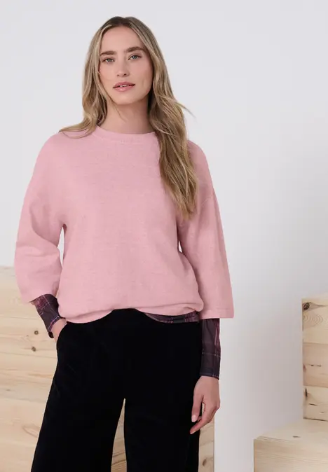 Junge Frau im rosa Pullover und schwarzen Hosen steht lässig mit Händen in den Taschen vor hellen Holzblöcken.