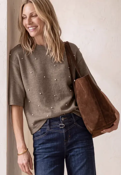 Une femme souriante en pull perlé, jean et sac en daim marron.