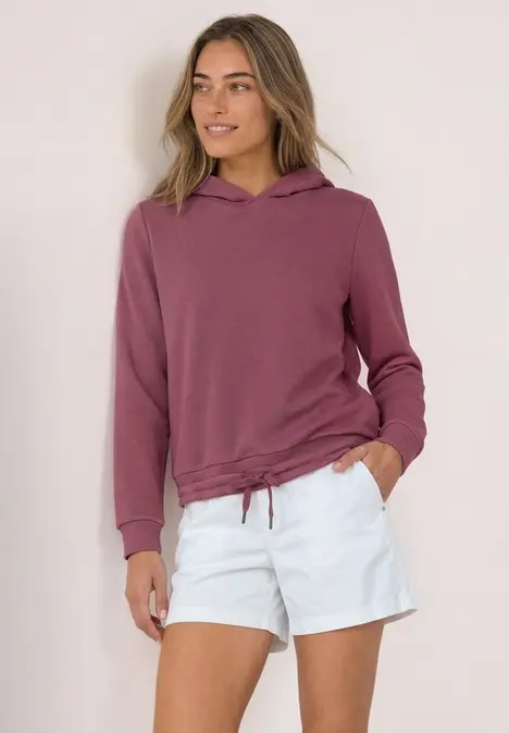 Frau in mauvefarbenem Hoodie mit Tunnelzug und weißen Shorts, steht entspannt mit Händen in den Taschen.