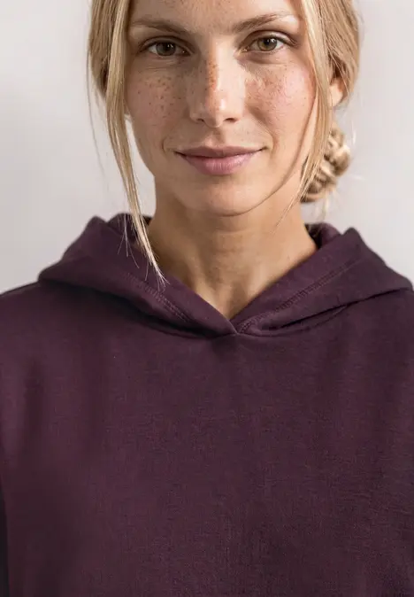 Nahaufnahme einer Frau mit Sommersprossen in bordeauxrotem Kapuzenpullover, Blick in die Kamera.