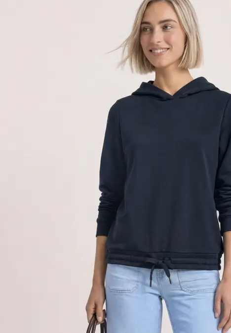 Eine lächelnde Frau in schwarzem Hoodie und hellblauen Jeans vor hellem Hintergrund.