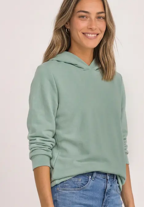 Frau in grünem Hoodie und Jeans lächelt in hellem Studio