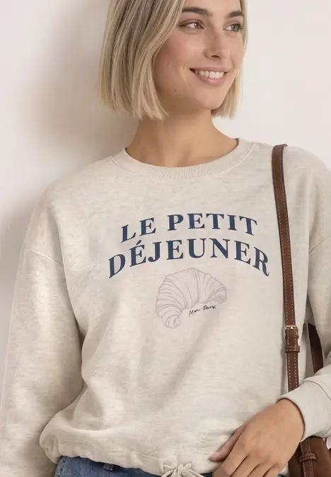 Frau trägt hellen Pullover mit Schriftzug «Le Petit Déjeuner» und Croissant-Illustration.