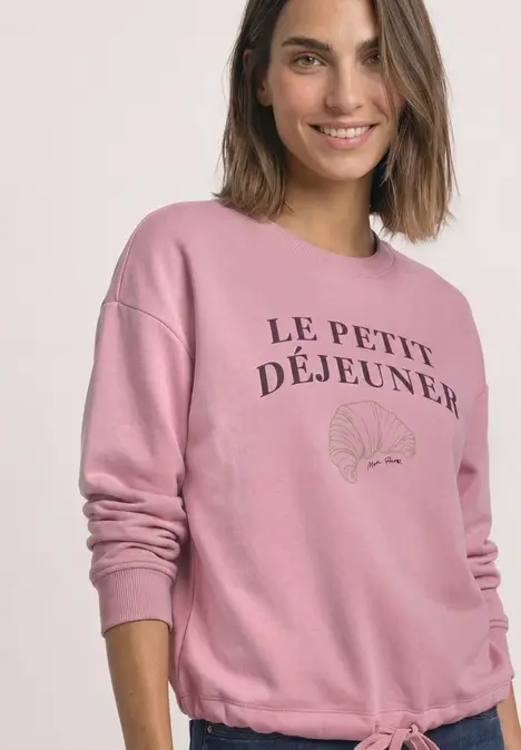 Frau trägt rosa Sweatshirt mit Aufschrift "Le Petit Déjeuner" und Croissant-Motiv, lächelt in die Kamera.