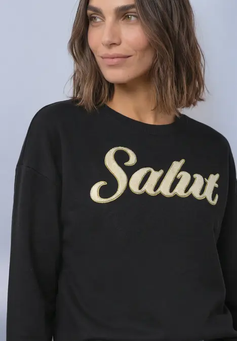 Schlichter schwarzer Pullover mit goldenem "Salut"-Schriftzug, getragen von einer lächelnden Frau.
