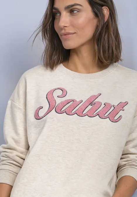 Frau in hellem Sweatshirt mit pinkem 'Salut'-Schriftzug, lächelnd vor schlichtem Hintergrund.