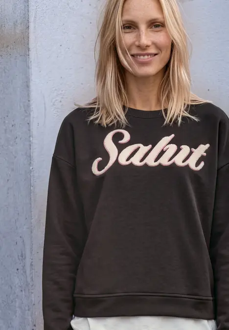 Fröhliche blonde Frau in schwarzem Pullover mit der Aufschrift 'Salut', lehnt lächelnd an einer Wand.