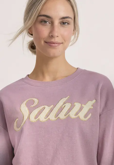 Junge Frau im rosa Sweatshirt mit goldener 'Salut'-Aufschrift lächelt dezent.