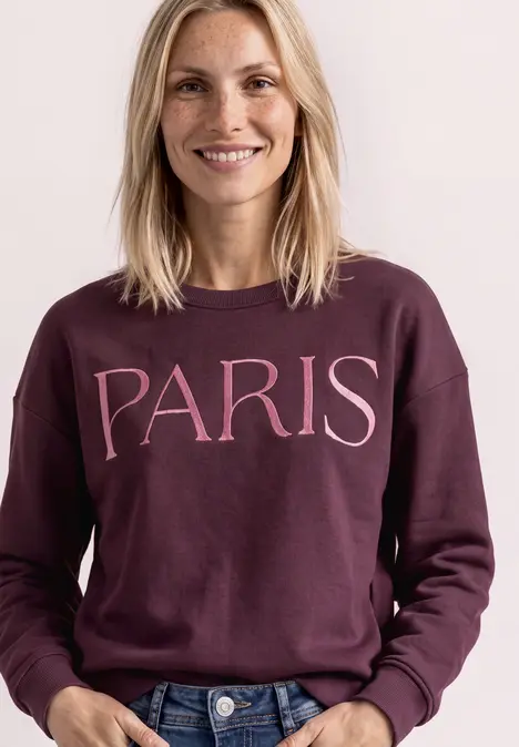 Vrouw lacht terwijl ze een bordeauxrode trui met 'PARIS' opdruk draagt.