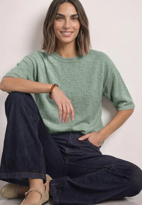 Frau mit grüner Bluse und dunkler Jeans sitzt entspannt und lächelt.