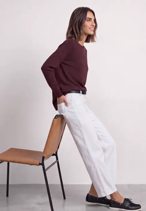 Vrouw in witte broek en bordeaux trui, leunt ontspannen tegen een stoel.
