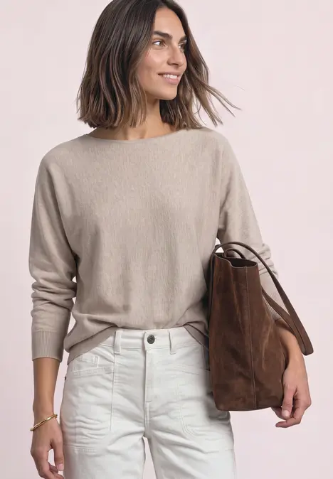 Frau mit schulterlangen Haaren, beigem Pullover, weißen Jeans und brauner Velours-Tasche.