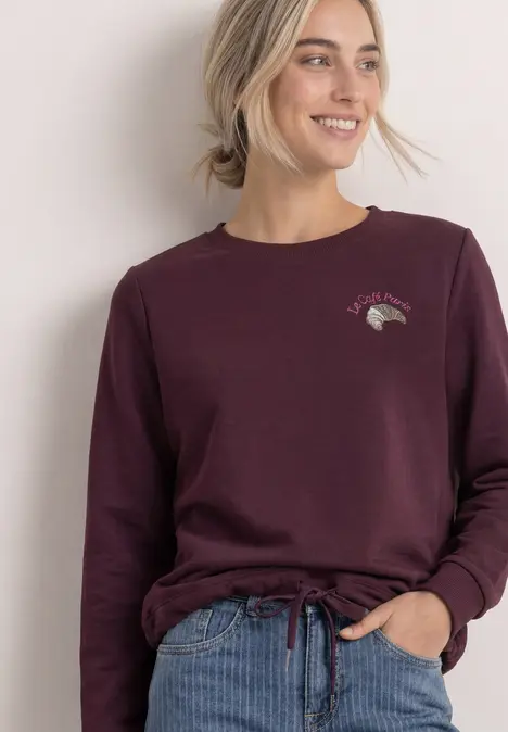 Frau in weinrotem Langarmshirt mit Tunnelzug, lächelnd vor hellem Hintergrund.
