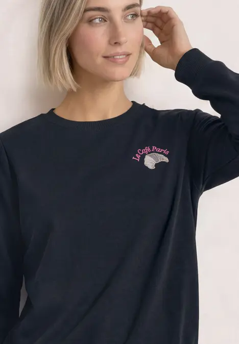 Frau mit kurzem blondem Haar in schwarzem Sweatshirt mit kleinem pink-grauen Logo auf der Brust.
