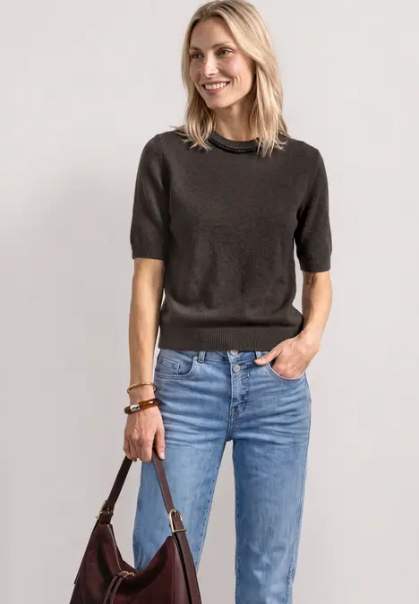 Lächelnde Frau in dunklem Kurzarmpullover, blauen Jeans und brauner Ledertasche.