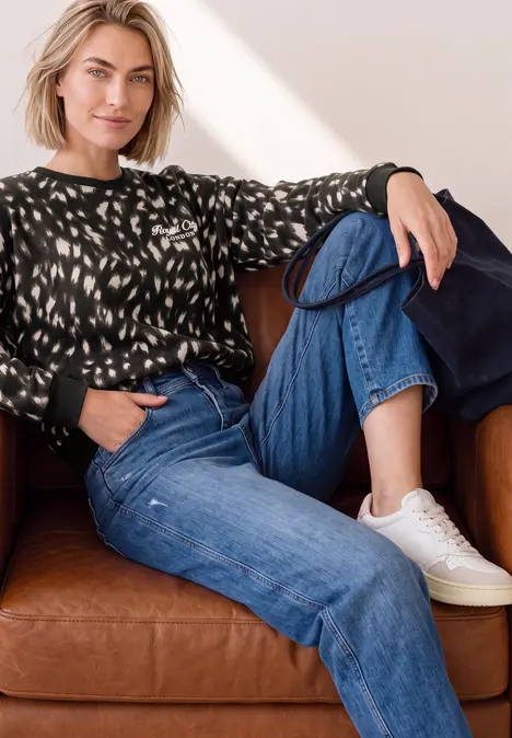 Eine Frau sitzt entspannt auf einem braunen Ledersofa, trägt ein gemustertes Sweatshirt, blaue Jeans und weiße Sneaker.