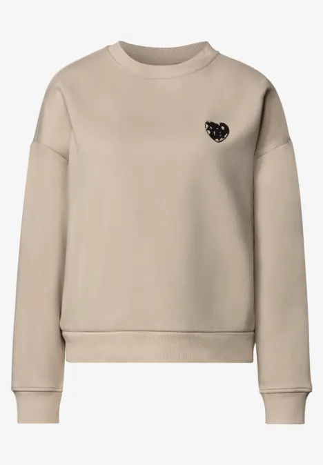 Beiges, schlichtes Sweatshirt mit Rundhalsausschnitt und kleinem schwarzen Herz-Emblem auf der Brust.