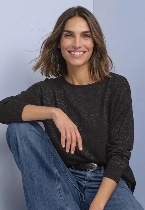 Eine lächelnde Frau mit braunen Haaren in dunkel meliertem Pullover und blauen Jeans sitzt entspannt.