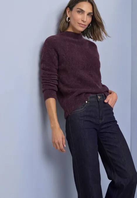 Frau lehnt an Wand, trägt weinroten Pullover und dunkle Jeans.