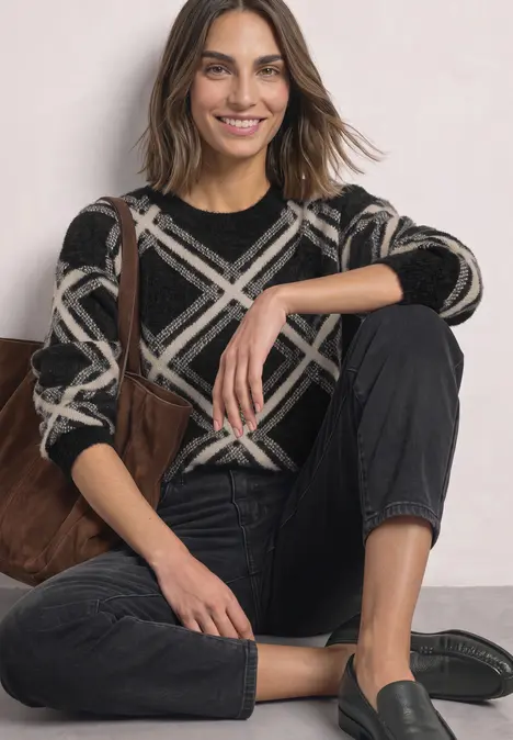 Frau sitzt lächelnd auf dem Boden, trägt schwarzen Pullover mit weißem Karomuster, schwarze Jeans und Loafers.
