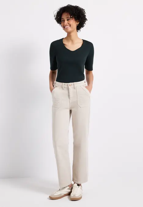 Frau mit dunklem Shirt, heller Hose und Sneakers lächelt mit Händen in den Taschen.