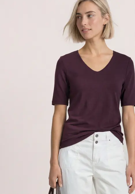 Frau mit kurzärmeligem, weinroten V-Shirt, lässig in weiße Jeans gesteckt auf hellem Hintergrund.