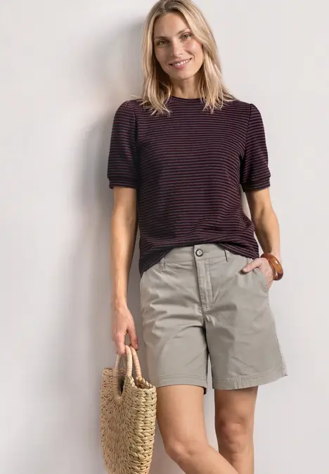 Frau in lila gestreiftem T-Shirt und grauen Shorts, hält eine Strohtasche, vor weißer Wand.