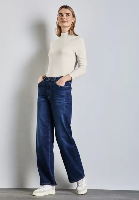 Frau in weißem Pullover und blauer Jeans steht mit Händen in den Taschen.