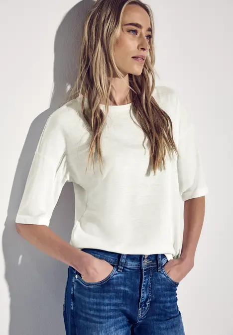 Frau in weißem Shirt und Jeans steht entspannt an der Wand.