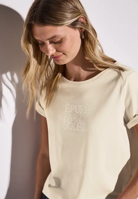 Frau mit blondem Haar trägt ein helles T-Shirt mit französischem Text.