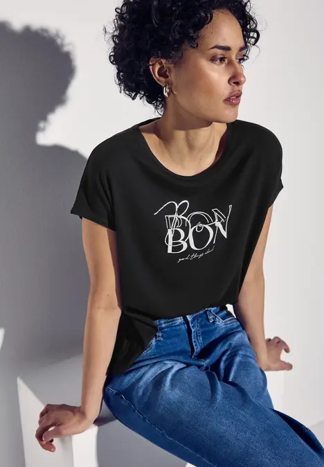 Frau mit lockigen Haaren trägt schwarzes T-Shirt mit Bonbon-Aufdruck und Jeans, sitzt entspannt.