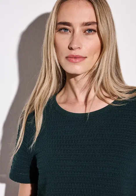 Frau mit langen blonden Haaren und dunklem Shirt vor weißer Wand.