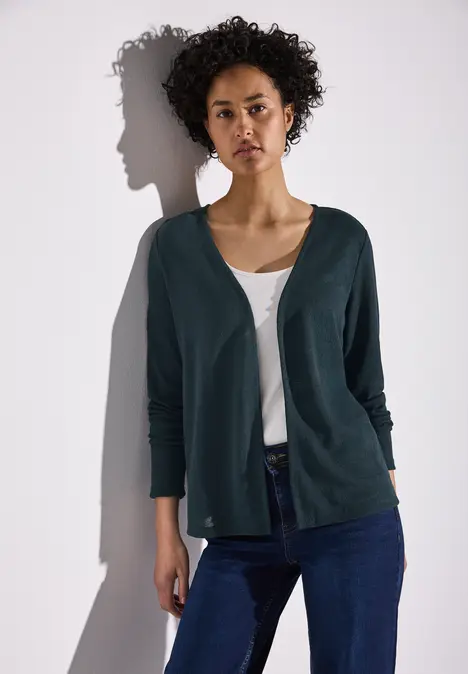 Frau mit lockigem Haar trägt dunkelgrüne Strickjacke, weißes Top und Jeans.