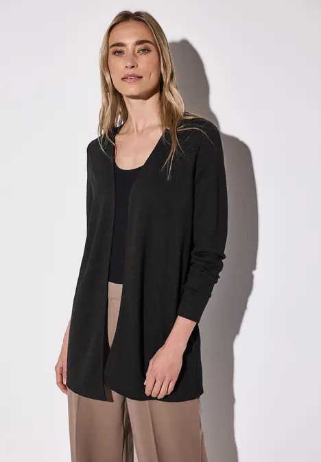 Frau mit schwarzem Cardigan, schwarzem Top und beigen Hosen vor hellem Hintergrund.