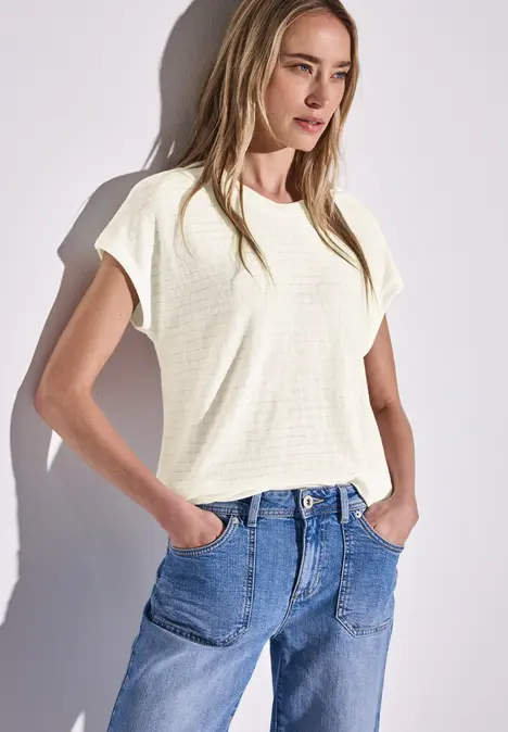 Frau mit langen Haaren trägt weißes T-Shirt und blaue Jeans, Hände in den Hosentaschen.