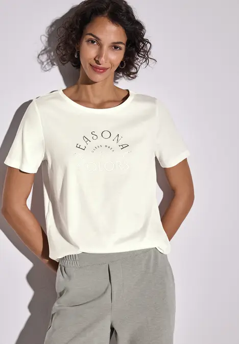 Frau mit lockigem Haar trägt weißes T-Shirt mit Aufdruck und graue Hose.