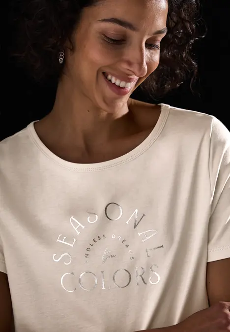 Lächelnde Frau mit lockigem Haar trägt ein Shirt mit silbernem Aufdruck.