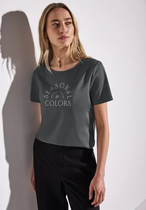 Frau mit dunklem T-Shirt, Aufschrift 'Seasonal Colors', steht vor weißer Wand.