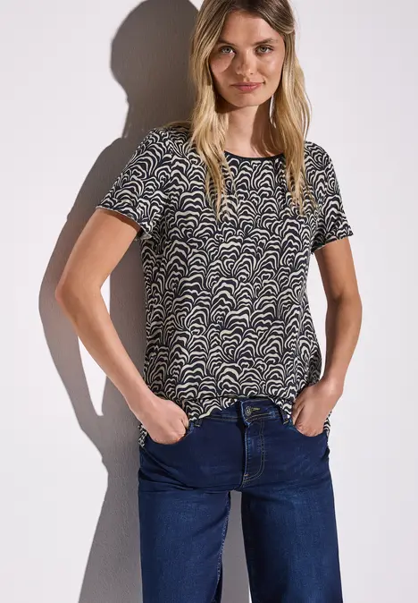 Frau mit gemustertem T-Shirt und Jeans, steht mit Händen in den Taschen vor weißer Wand.